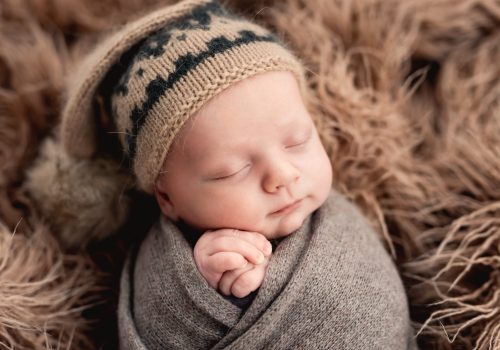 Newborn baby boy swaddled in fabric sleeping and holding his hands close to cheeks. Infant kid napping on fur